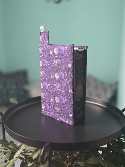 Couvre-livre artisanal ajustable aux nombres de pages et à l'épaisseur du livre. Motif tissu en coton chat  ésotérique, lune d'améthyste avec son marque-page en tissu assorti. Posé sur une table noire.
