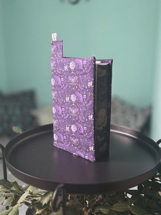 Couvre-livre artisanal ajustable aux nombres de pages et à l'épaisseur du livre. Motif tissu en coton chat  ésotérique, lune d'améthyste avec son marque-page en tissu assorti. Posé sur une table noire.