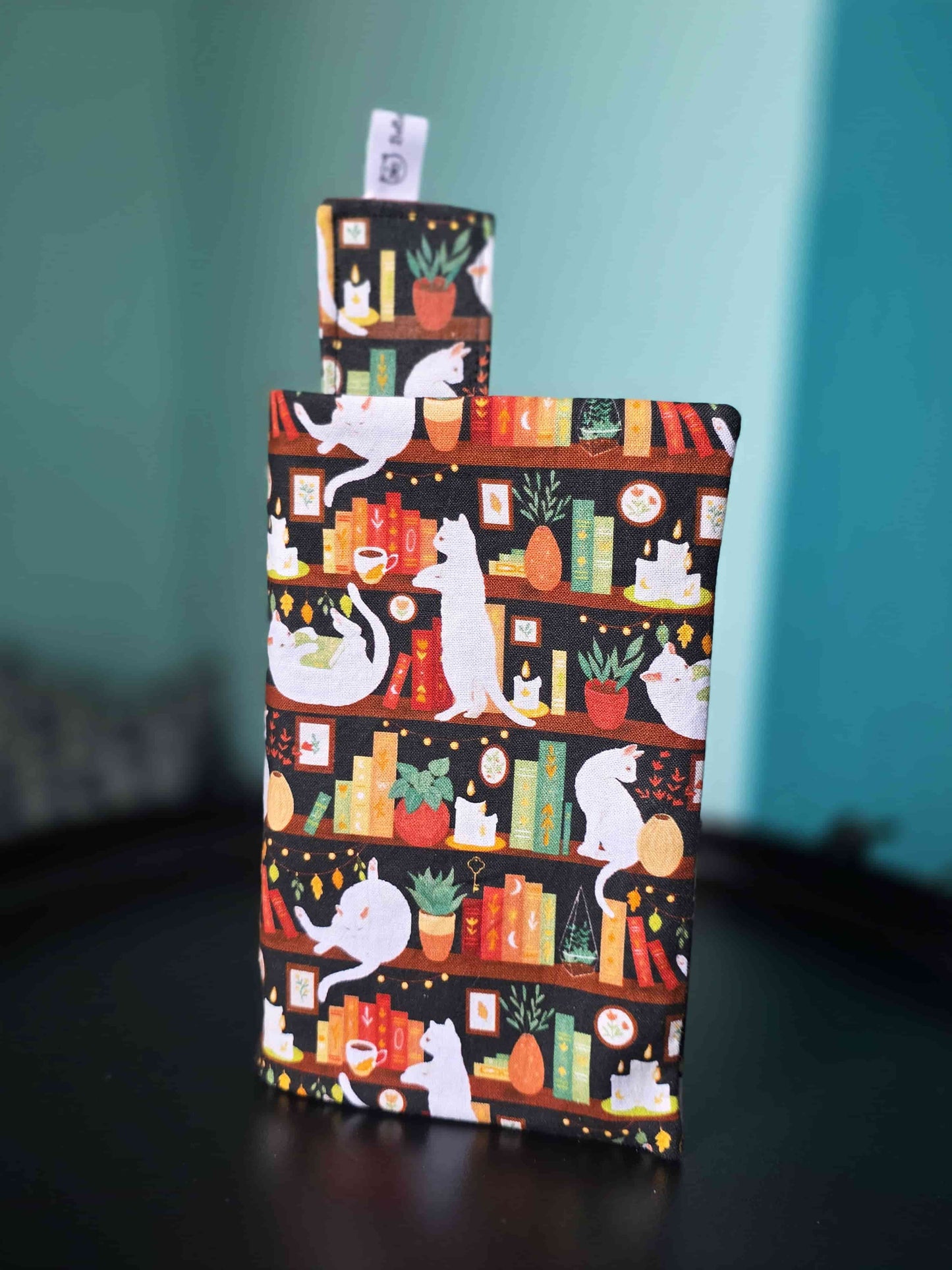 Couvre-livre artisanal ajustable aux nombres de pages et à l'épaisseur du livre. Motif tissu en coton chat bibliothèque avec son marque-page en tissu assorti. Posé sur une table noire.