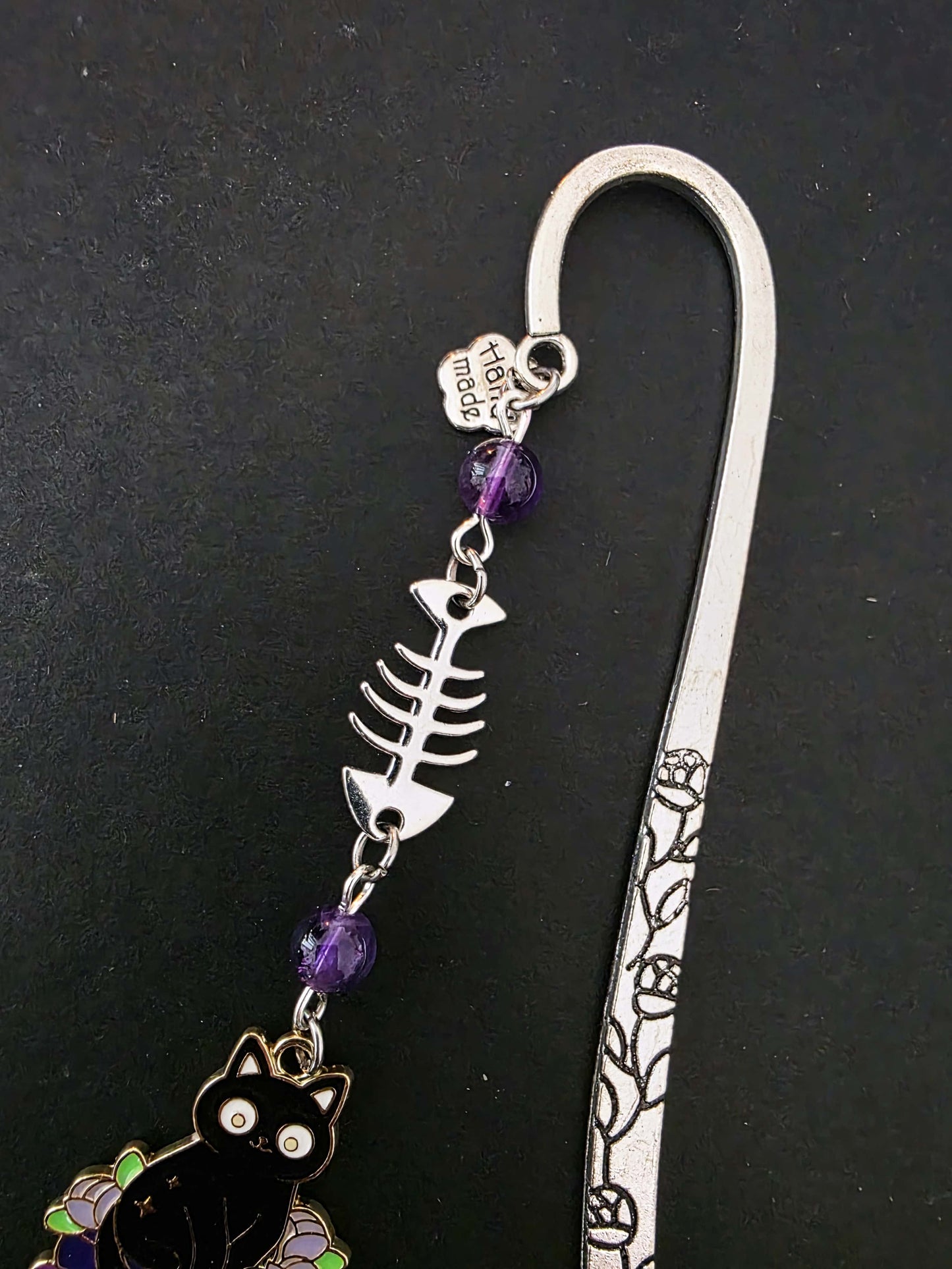 Marque-page en métal d'un chat noir assis sur une pile de livres violets évoquant des grimoire. Une arête de poisson est en breloque au centre et il y a deux pierres naturelles d'améthyste de chaque côté. Le bijou de livre est posé sur un livre noir 