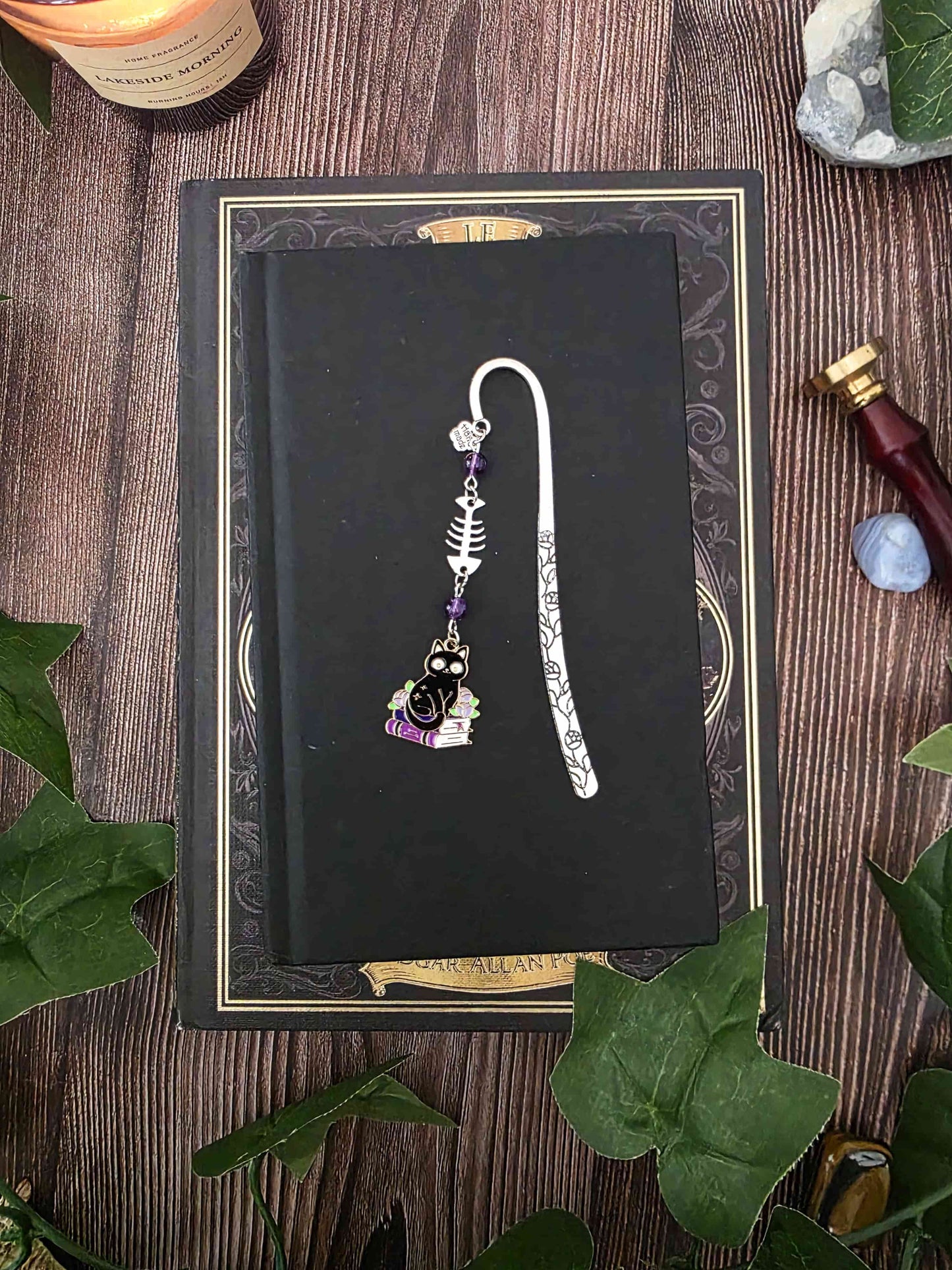 Marque-page en métal d'un chat noir assis sur une pile de livres violets évoquant des grimoire. Une arête de poisson est en breloque au centre et il y a deux pierres naturelles d'améthyste de chaque côté. Le bijou de livre est posé sur un livre noir 