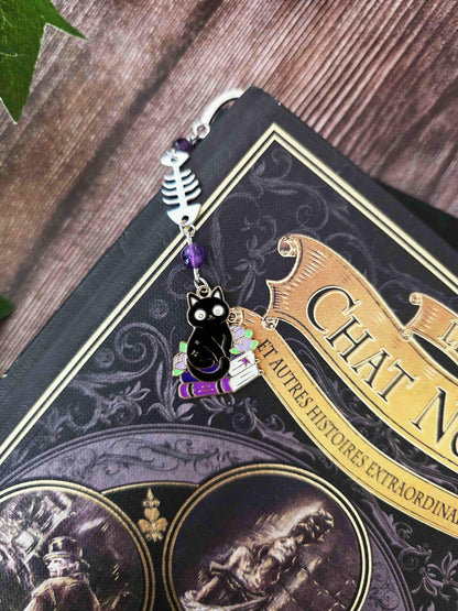 Marque-page en métal d'un chat noir assis sur une pile de livres violets évoquant des grimoire. Une arête de poisson est en breloque au centre et il y a deux pierres naturelles d'améthyste de chaque côté. Le bijou de livre est posé sur un livre de Edgar allan poe