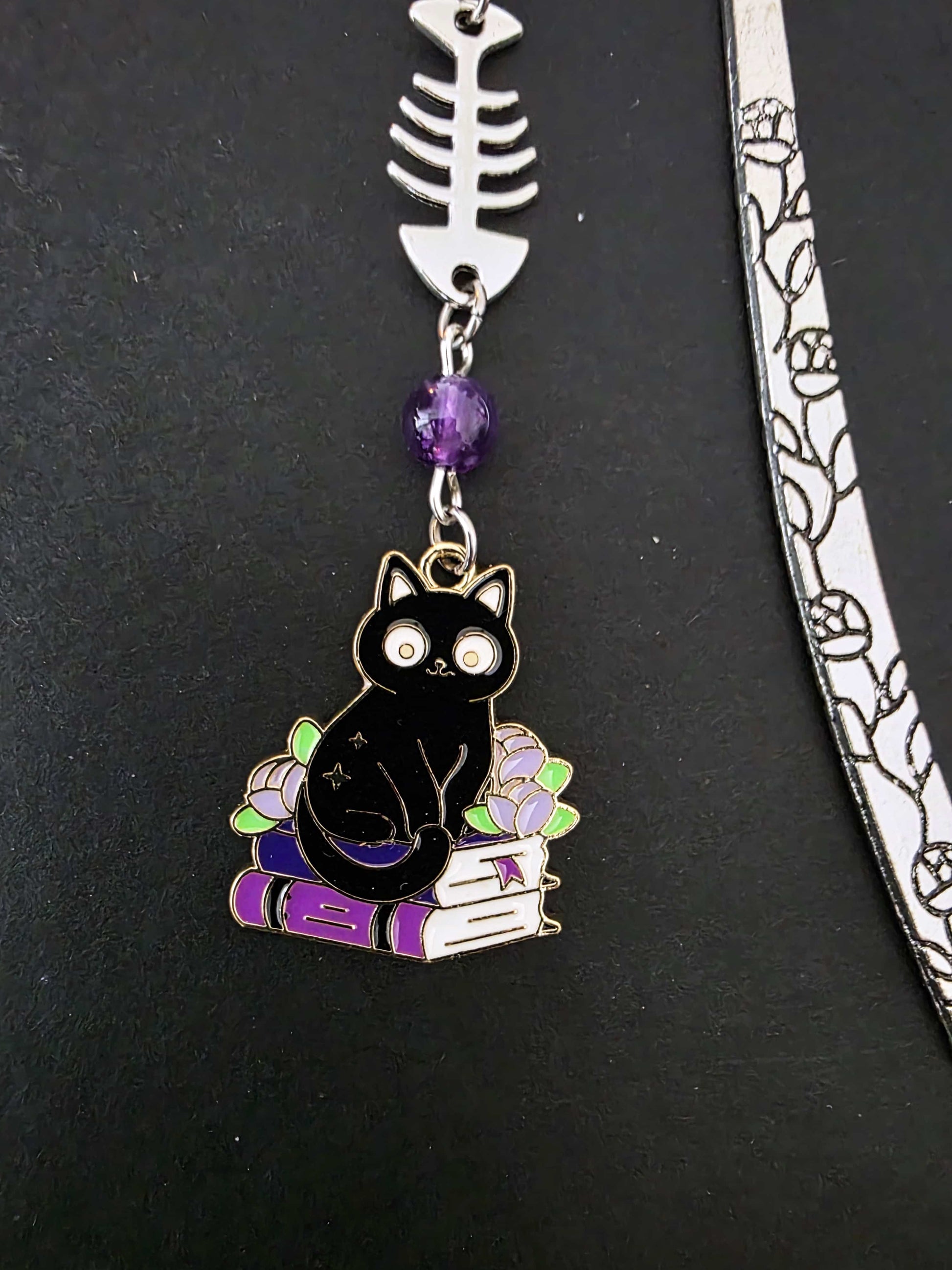 Marque-page en métal d'un chat noir assis sur une pile de livres violets évoquant des grimoire. Une arête de poisson est en breloque au centre et il y a deux pierres naturelles d'améthyste de chaque côté. Le bijou de livre est posé sur un livre noir 