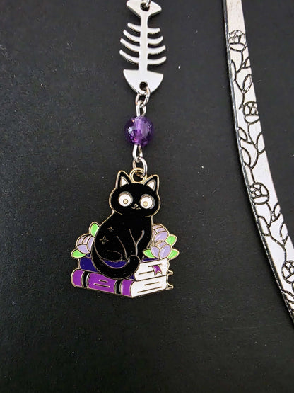 Marque-page en métal d'un chat noir assis sur une pile de livres violets évoquant des grimoire. Une arête de poisson est en breloque au centre et il y a deux pierres naturelles d'améthyste de chaque côté. Le bijou de livre est posé sur un livre noir 