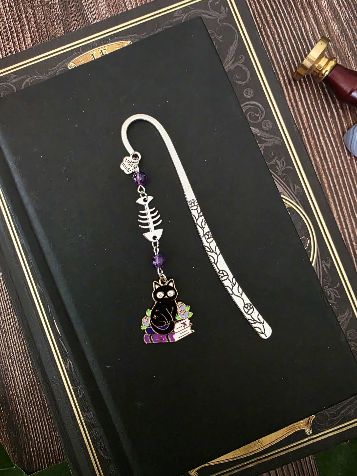 Marque-page en métal d'un chat noir assis sur une pile de livres violets évoquant des grimoire. Une arête de poisson est en breloque au centre et il y a deux pierres naturelles d'améthyste de chaque côté. Le bijou de livre est posé sur un livre noir 