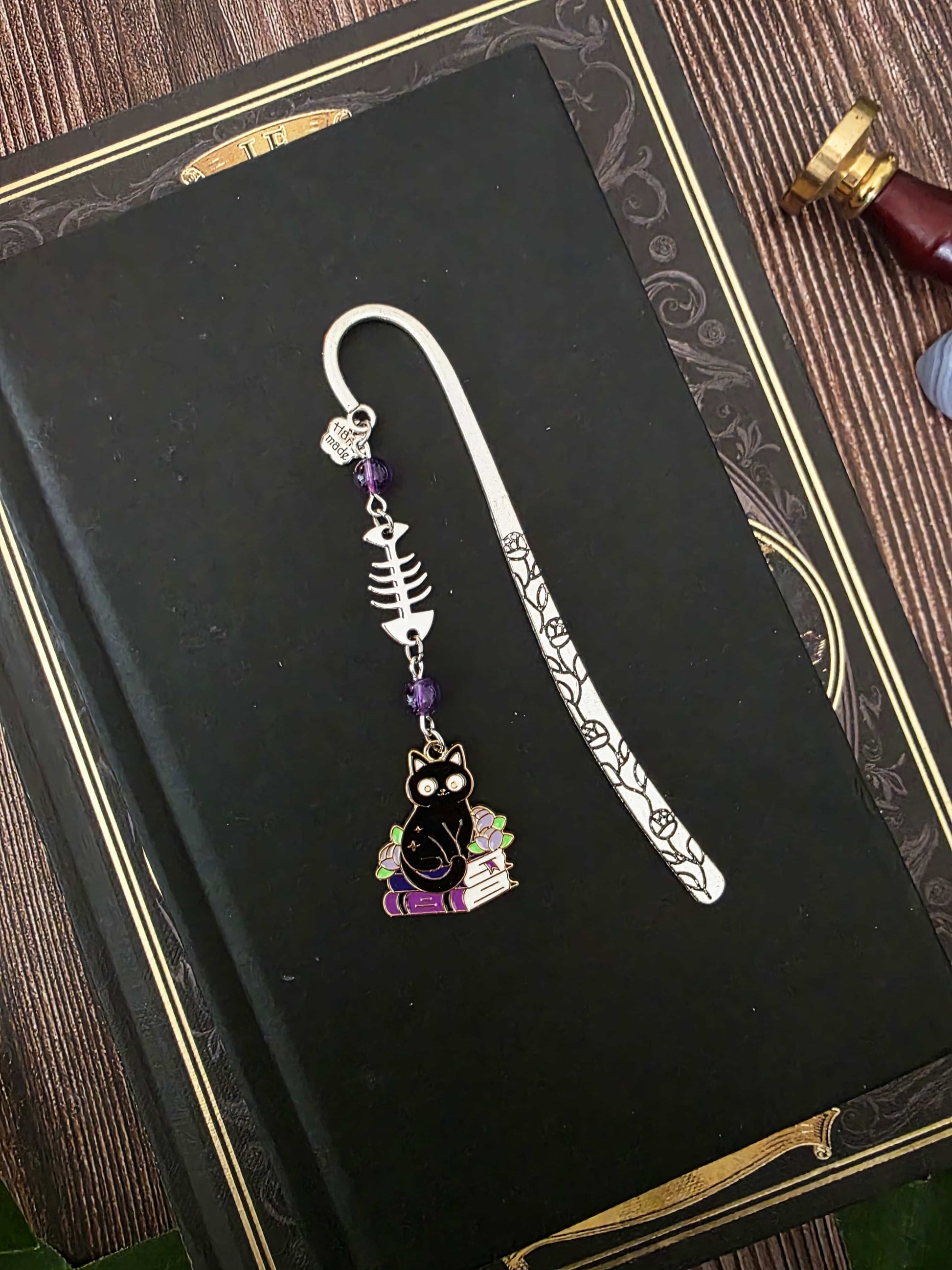 Marque-page en métal d'un chat noir assis sur une pile de livres violets évoquant des grimoire. Une arête de poisson est en breloque au centre et il y a deux pierres naturelles d'améthyste de chaque côté. Le bijou de livre est posé sur un livre noir 