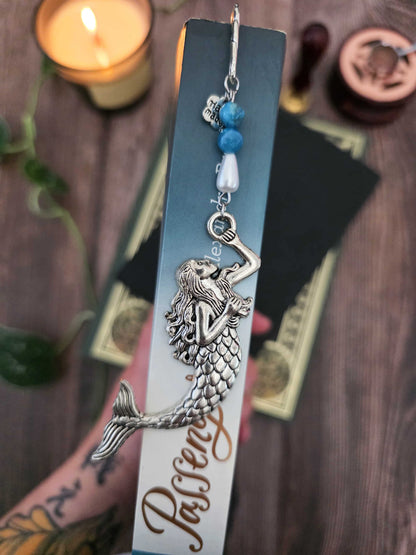 marque page en metal, signet sirene avec deux perles d'apatite bleue et une perle en forme de goutte blanche. Sur une tranche de livre "Passenger" sur la mer.